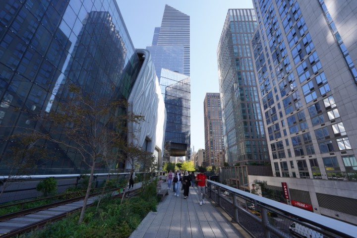 A section of the High Line pathway in NYC. Many people are walking on the trail. Vegetation is visible along the trail. And tall buildings stand on either side.